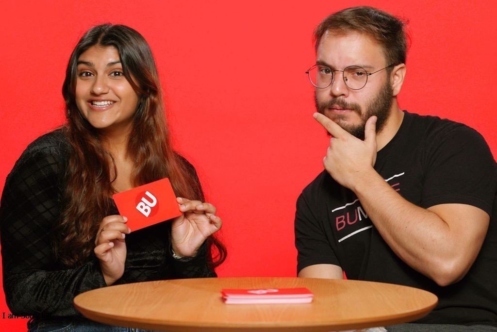 Photo: A picture of two students, a girl on the left and a boy on the right, posing in front of a red background. They are sitting at a table while the girl smiles and holds a red card that says "BU" and the boy places his pointer finger and thumb against his chin.